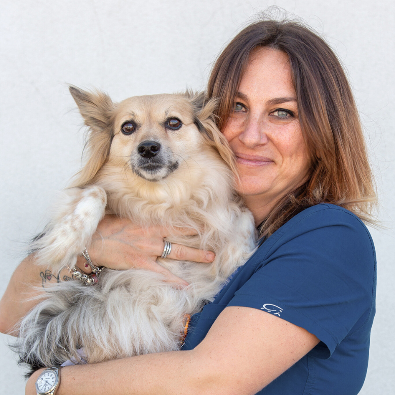 13 Una donna in camice blu sorride mentre tiene in braccio un soffice cane bianco e abbronzato. La donna ha capelli castani lunghi fino alle spalle e gioielli al polso, che incarnano la cura dei Servizi Veterinari. Entrambi appaiono soddisfatti su uno sfondo chiaro.