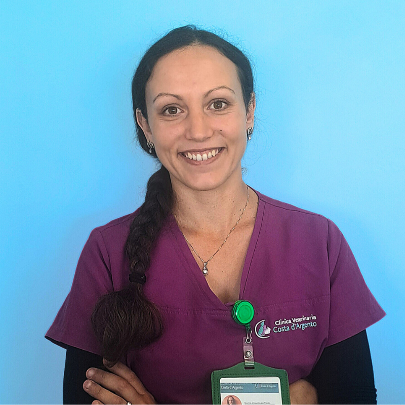Claudia Loffredo front office della Clinica Veterinaria Costa d'Argento. Una donna con lunghi capelli castani raccolti in una treccia, che indossa un camice viola e un distintivo della clinica, sta sorridendo con le braccia incrociate davanti a uno sfondo azzurro.