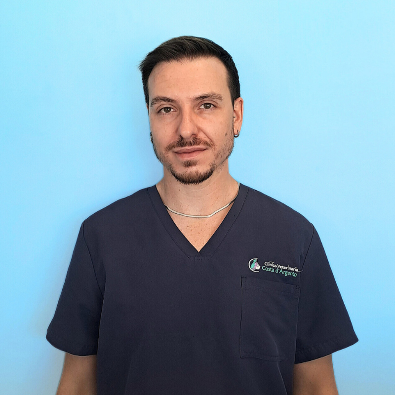 Il Dottor Manuele Sabatini, medico veterinario della Clinica Veterinaria Costa d'Argento. Un uomo con capelli corti e scuri e barba curata, che indossa un camice da medico blu navy, è in piedi su uno sfondo blu chiaro.