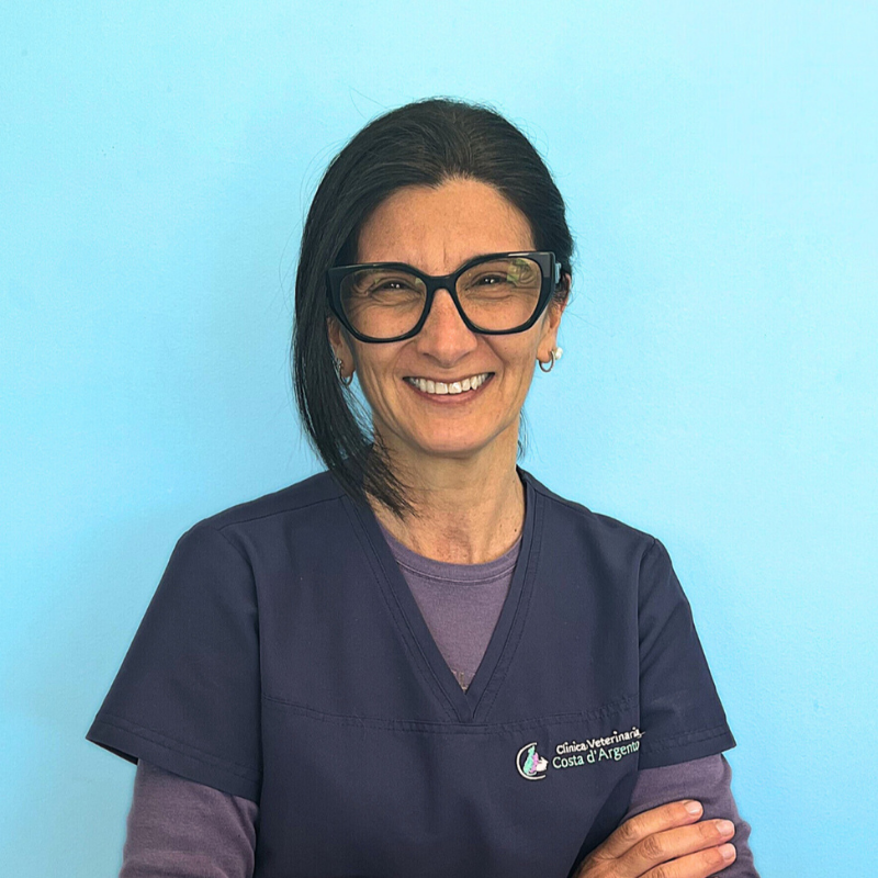 La Dottoressa Sandra Capelloni, co-fondatrice e direttore sanitario della Clinica Veterinaria Costa d'Argento. Una donna dai lunghi capelli scuri, con occhiali e camice medico blu navy, è in piedi sorridente con le braccia incrociate davanti a uno sfondo azzurro.