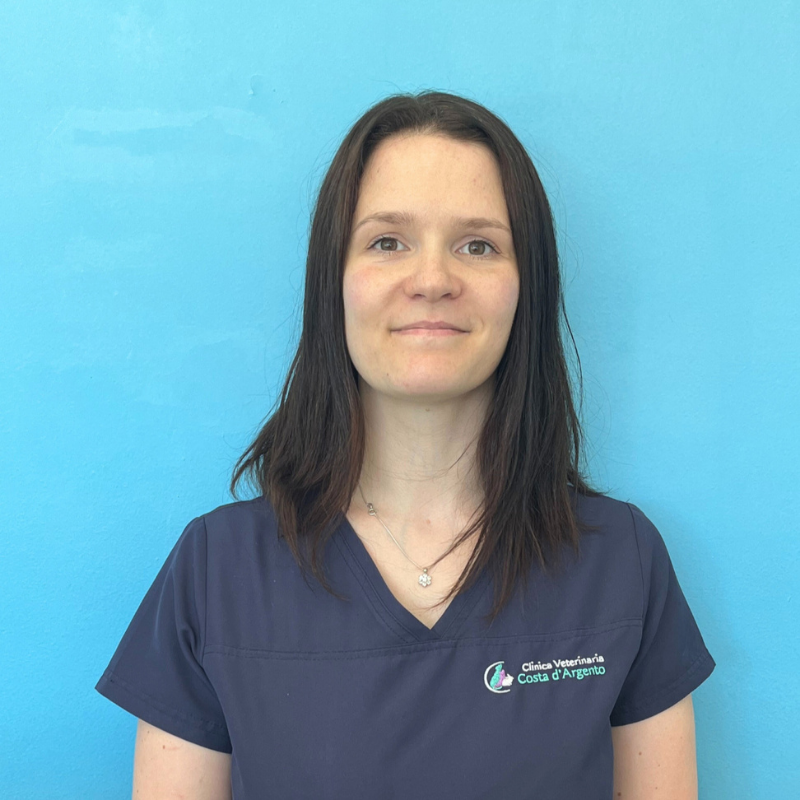 La dottoressa Silvia Mazzolai, medico veterinario della Clinica Veterinaria Costa d'Argento. Una donna con i capelli lisci e castani che indossa un camice blu navy è in piedi di fronte a una parete blu brillante, guarda la telecamera e sorride leggermente. Sul camice c'è un logo.