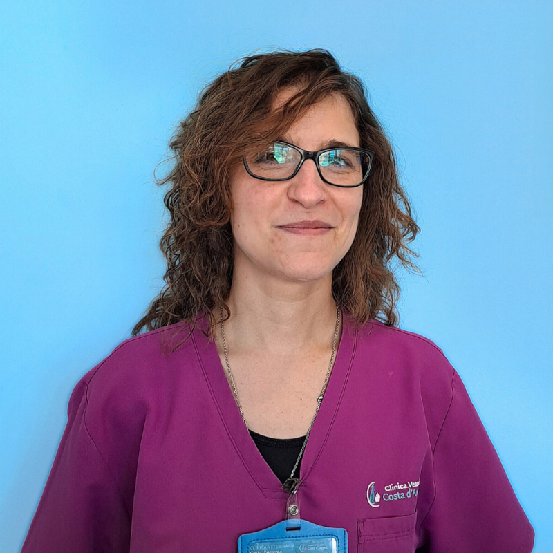 Laura Graziani. back office della Clinica Veterinaria Costa d'Argento. Una donna con capelli castani ondulati e occhiali, che indossa un camice da medico viola e un distintivo, si trova davanti a uno sfondo azzurro, sorridendo leggermente.