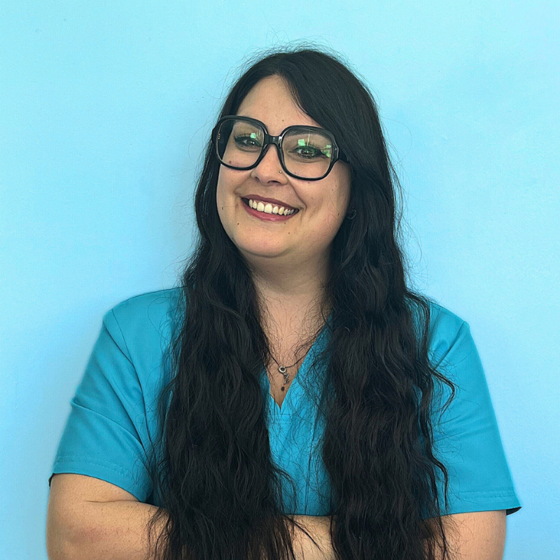 Valentina Bellini, tecnico veterinario della Clinica Veterinaria Costa d'Argento. Una donna sorridente con lunghi capelli scuri e occhiali, che indossa un camice verde acqua, si trova su uno sfondo azzurro con le braccia incrociate.